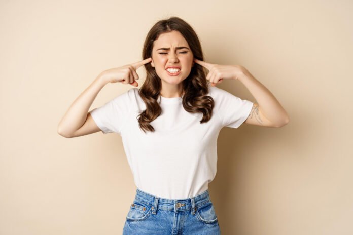 annoyed young woman shut ears from loud noise feeling discomfort complaining standing against beige background